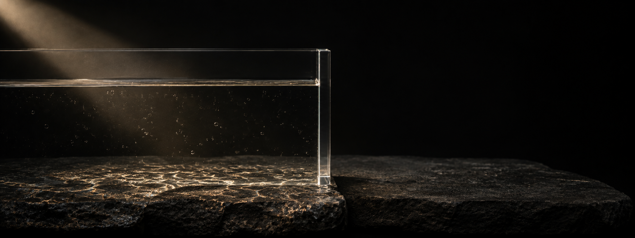 A single flawless slab of machined glass standing in dark stone. Water is held perfectly level against one face of it; the stone on the other side is dry and unlit. The light stops at the glass.